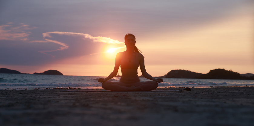 Eine Frau sitzt in der typische Yogaposition am Strand beim Sonnenuntergang